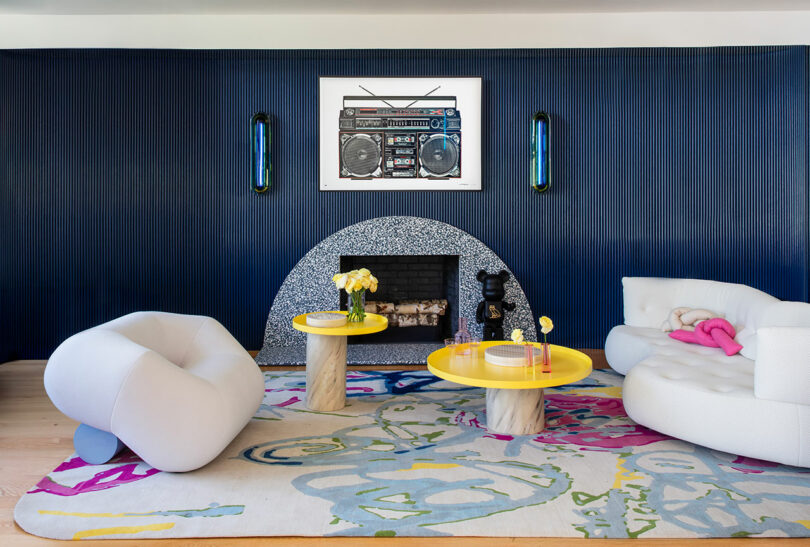 Modern living room with blue paneled walls, terrazzo fireplace, white sculptural chairs, yellow tables, abstract rug, and framed boombox art above the mantle.