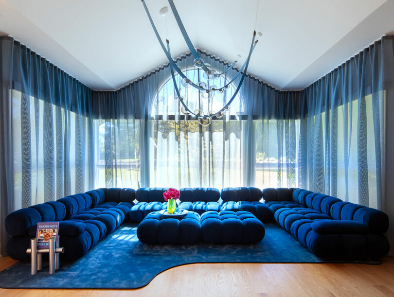 A modern living room with a large blue sectional sofa, blue rug, glass coffee table with pink flowers, floor-to-ceiling sheer blue curtains, and a contemporary chandelier.