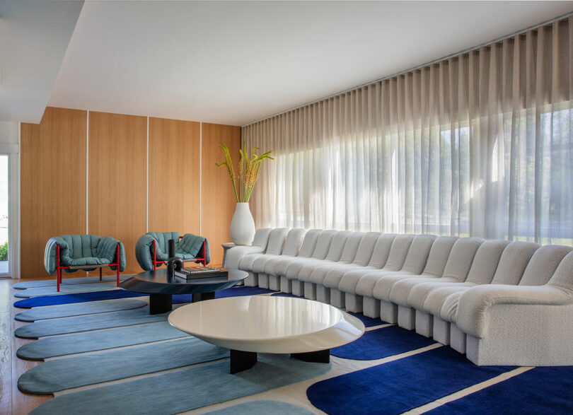 Modern living room with a long white sectional sofa, two teal armchairs, large white vase with tall plants, round coffee tables, and blue patterned rug. Floor-to-ceiling windows with sheer curtains.