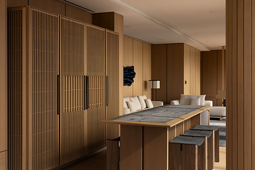 Modern kitchen and living area with wood paneling, a long marble-top island with stools, and a white sofa set in the background.
