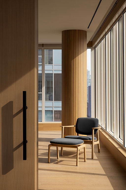 A minimalist lounge area with a wooden chair and ottoman next to large windows, a wooden column, and natural light filling the space.