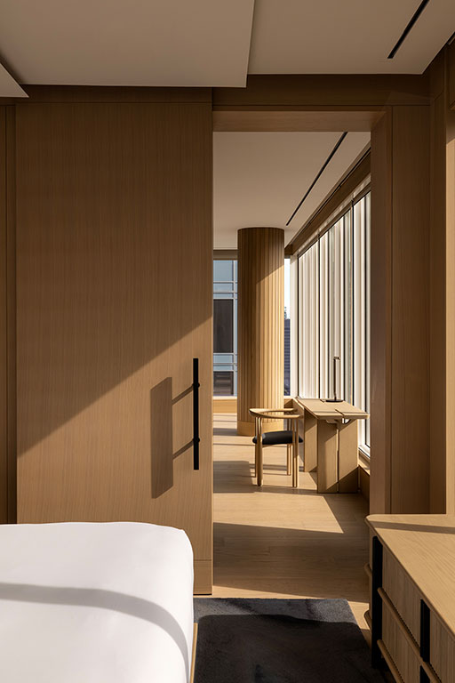 A minimalist bedroom with light wood walls and furniture, large windows, a bed in the foreground, and a desk with a chair in the sunlit room beyond.