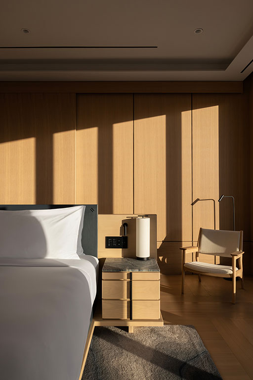 A modern hotel room with a neatly made bed, a nightstand holding a kettle, a wooden chair, and sunlight casting shadows on wood-paneled walls.