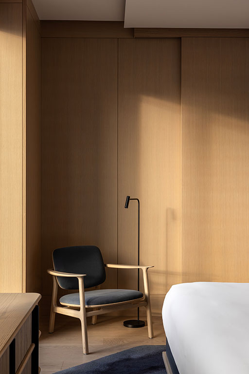 A modern bedroom corner with a wooden armchair with dark cushions, a black floor lamp, and light wood paneling, partially lit by natural light.