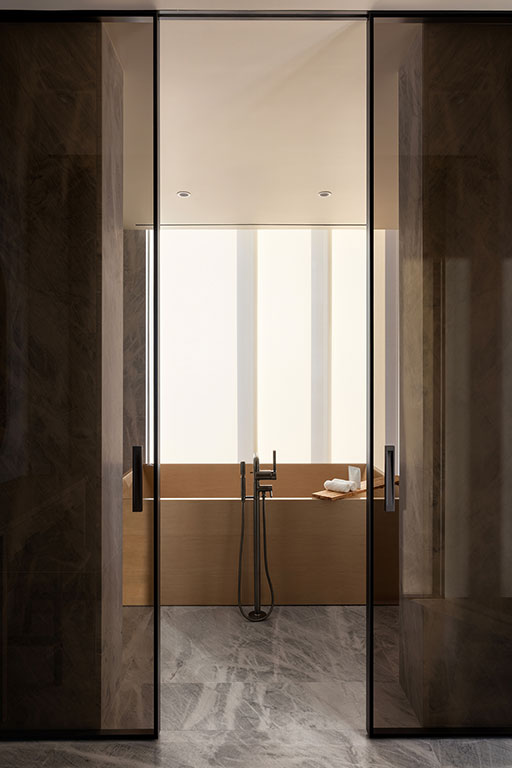 A modern bathroom with a wooden bathtub, floor-mounted faucet, towel, and natural light from tall frosted windows, viewed through glass doors.