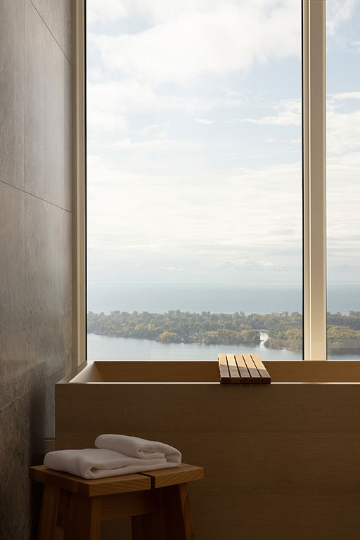 A modern bathtub positioned by a large window with a view of a lake and distant shoreline; folded white towels rest on a wooden stool nearby.