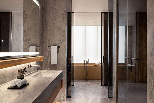 Modern bathroom with marble surfaces, a large mirror, a sink with rolled towels, glass shower doors, and a freestanding bathtub near a frosted window.