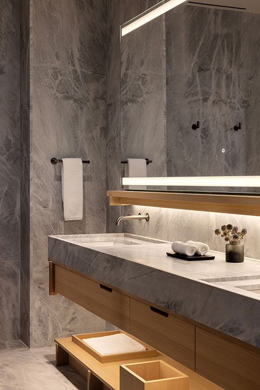 Modern bathroom with gray marble walls and countertop, large lit mirror, wooden vanity with open drawers, towels, and decorative flowers on the counter.