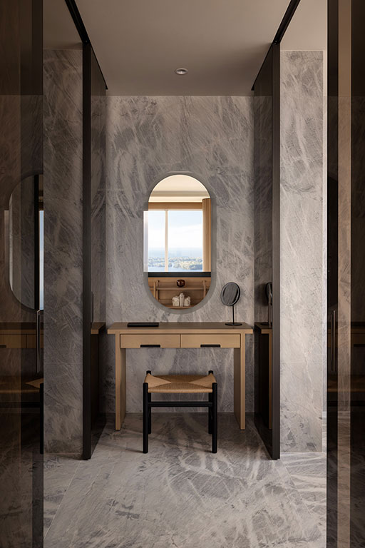A minimalist vanity area with a wooden table, round mirror, small stool, and marble walls and floor, with a window and city view in the background.