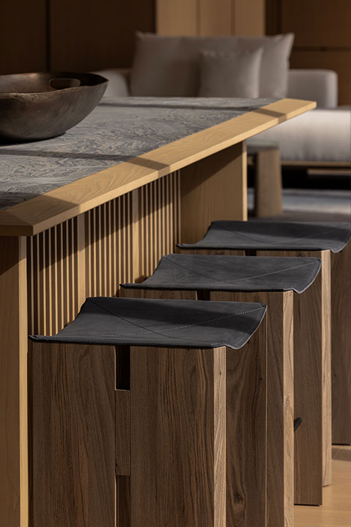 A row of wooden bar stools with black seats is arranged along a kitchen island with a gray stone countertop and wooden paneling.