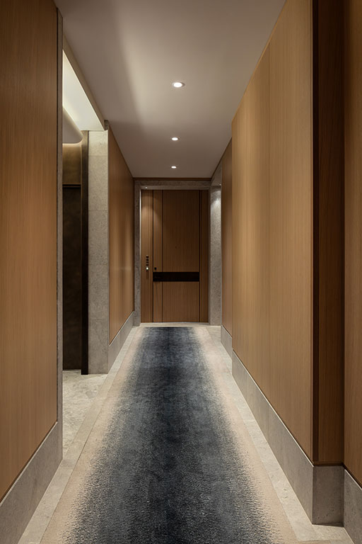 A modern hallway with wood-paneled walls, recessed lighting, a dark carpet runner, and a closed door at the end.