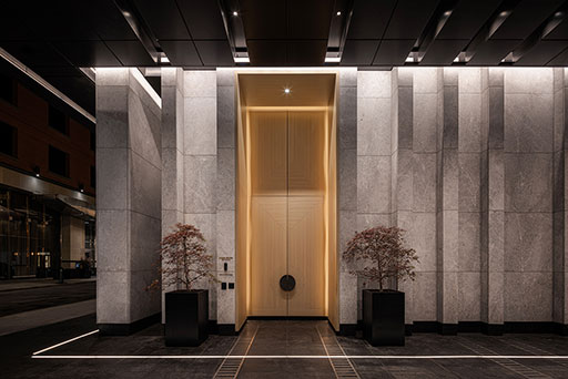 A modern building entrance with a tall illuminated wooden door, flanked by two potted trees, surrounded by gray stone walls at night.