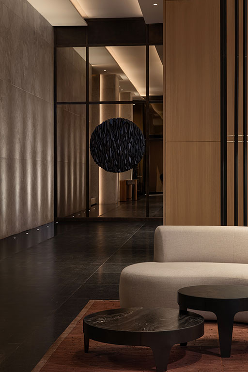 Modern interior with beige sofa, black round coffee tables, wood and marble wall panels, and a sculptural black sphere suspended in front of a mirrored wall.
