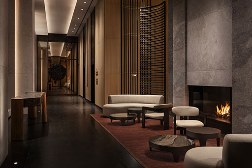 Modern hotel lobby with minimalist furniture, a lit fireplace, stone walls, dark flooring, and warm lighting. A hallway with a decorative art piece is visible in the background.