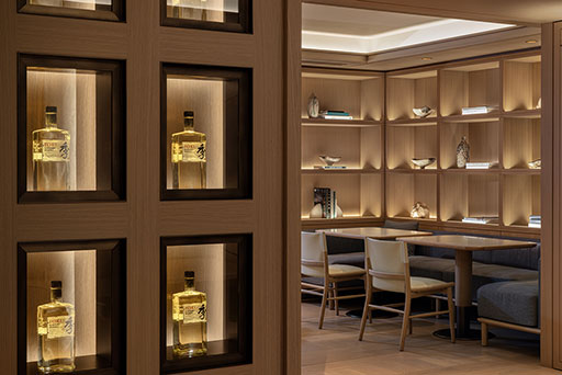 Backlit shelves display bottles of whiskey on a wooden wall; in the background, a modern lounge area features tables, chairs, and decorative shelves.