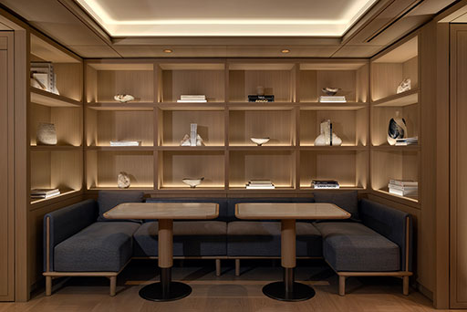 A modern lounge with two wooden tables, dark cushioned benches, and built-in shelves displaying books and decorative objects, all under warm lighting.