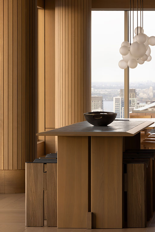 Modern dining area with a wooden table, dark bowl centerpiece, wooden stools, paneled walls, large window, and cluster of white pendant lights. Cityscape visible outside.