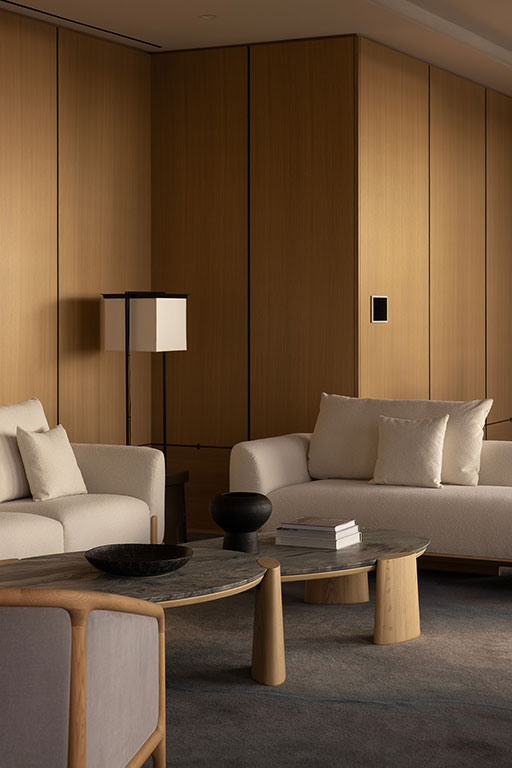 Minimalist living room with light wood-paneled walls, two white sofas, a square floor lamp, and a low coffee table holding a black bowl and tray.