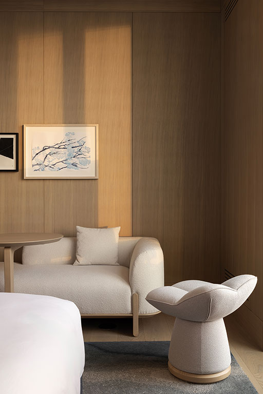 A light-colored modern armchair and side table sit against a wooden wall with framed art, illuminated by natural light.