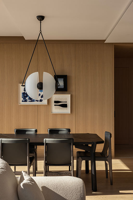 A modern dining area with a black table, five black chairs, a sculptural pendant light, wood-paneled walls, and framed abstract artwork.