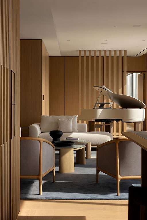 Modern living room with light wood paneling, minimalist furniture, a gray area rug, and a white grand piano near vertical wooden slats.