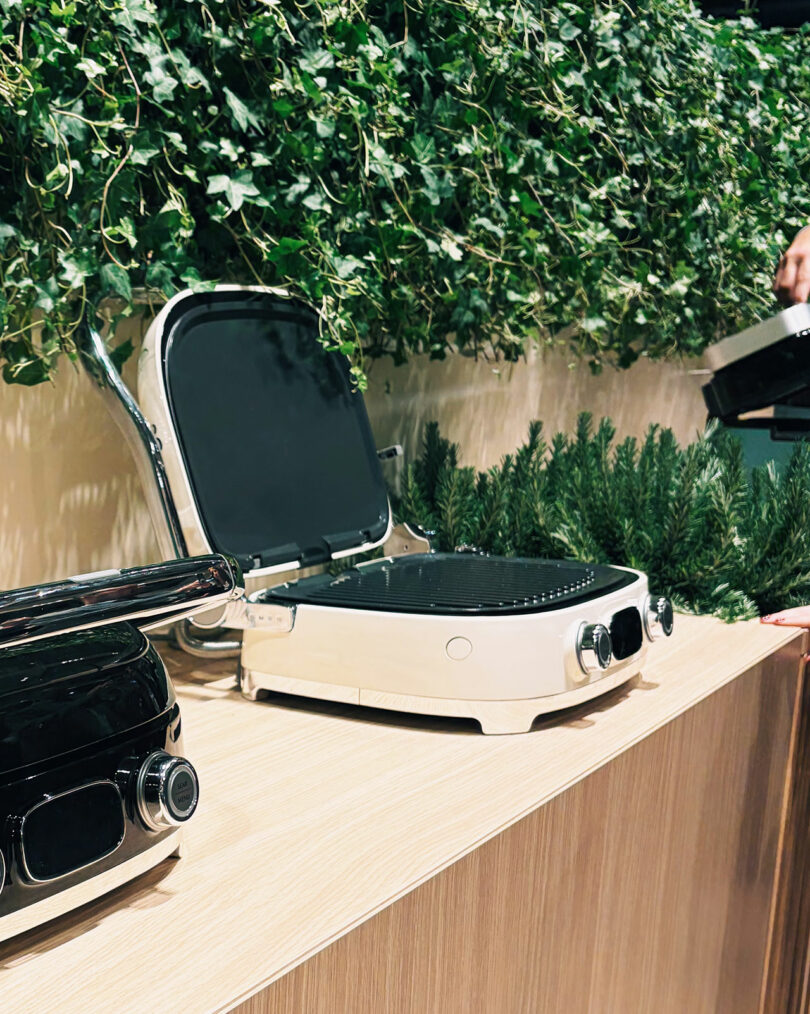 Two electric grills, one black and one white, sit on a wooden counter with green plants in the background; a person holds a grill lid on the right side of the image.