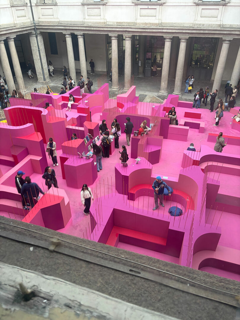 Aerial view of people walking through a large, bright pink maze installation in an open courtyard surrounded by columns.