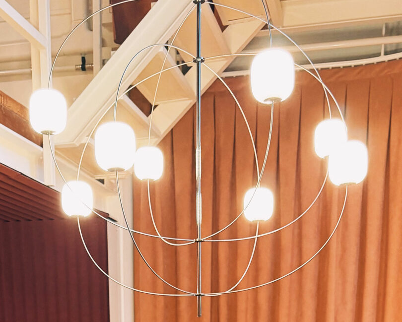Modern chandelier with eight glowing white glass globes and intersecting metal rings, suspended from the ceiling in a room with brown curtains.