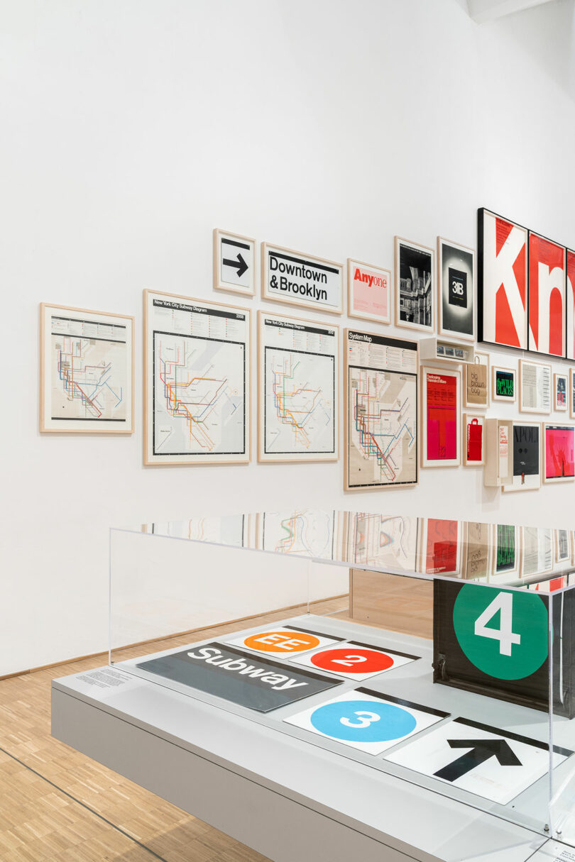 Wall-mounted subway maps and transit signs are displayed alongside framed posters in a modern gallery. A glass case showcases additional transit signage on a table.