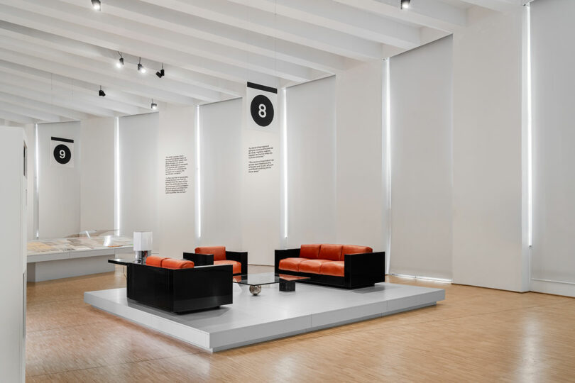 A modern exhibition room with white walls, orange and black sofas on a platform, and display signs numbered 8 and 9 hanging from the ceiling.
