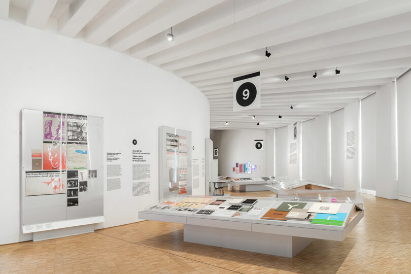 Modern museum exhibition space with white walls, display cases of documents and objects, wood flooring, and numbered black signs hanging from the ceiling.