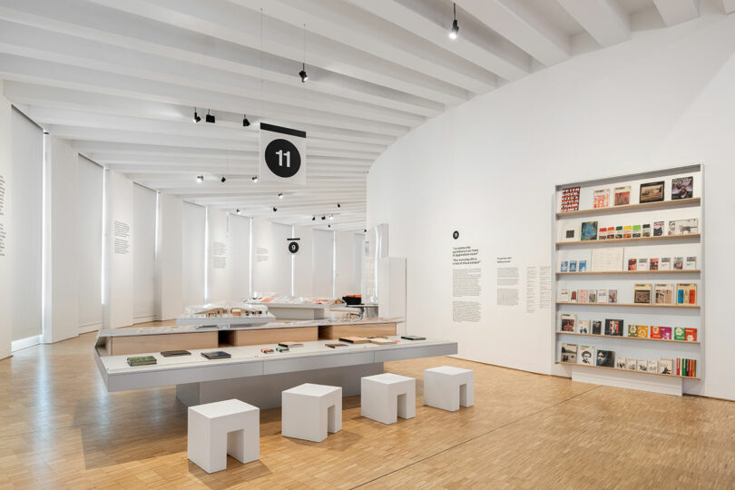 A modern, minimalist museum exhibit with books and magazines displayed on tables and shelves, numbered section 11, with white walls, wooden floor, and simple white stools.