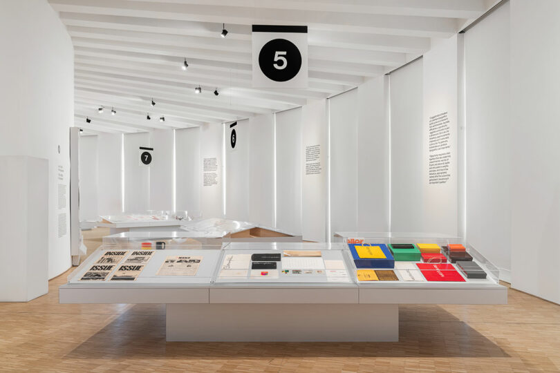 A modern museum exhibit displays books, magazines, and colorful objects under glass, with white walls, wood flooring, and numbered signs hanging from the ceiling.