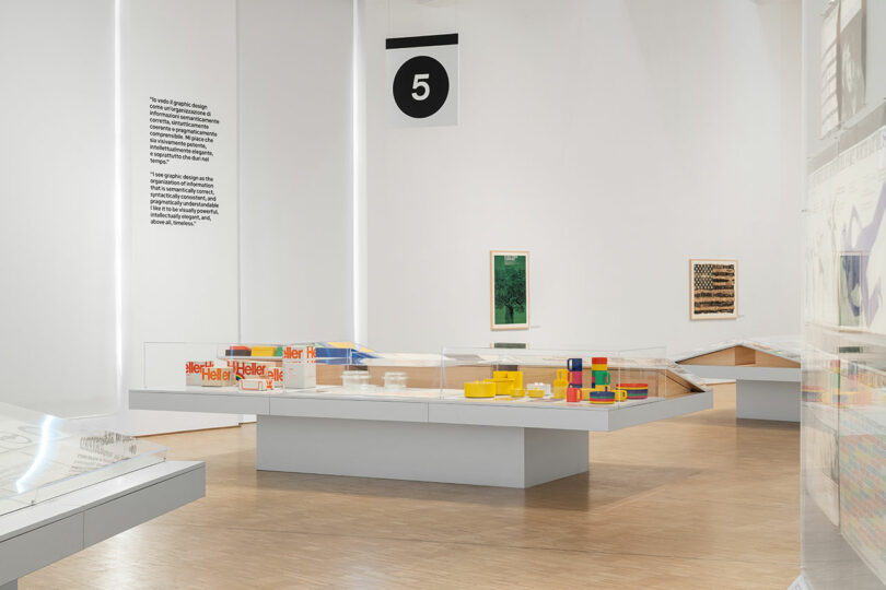A modern museum exhibit with display cases containing colorful objects, wall text, framed artwork, and a large black and white number 5 sign hanging above.