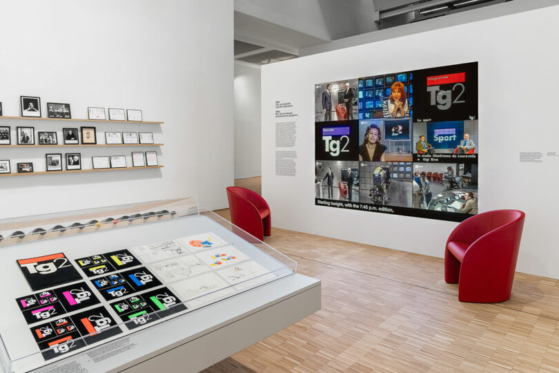 Exhibition room with display cases of graphic designs, framed images on the wall, two red chairs, and a large wall projection featuring various broadcast images and logos.