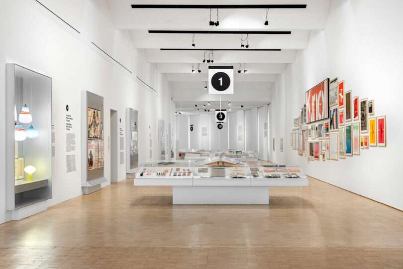 A modern museum gallery with white walls, display cases, graphic posters on the walls, and hanging signs marked with numbers; hardwood floors and ceiling lights are visible.