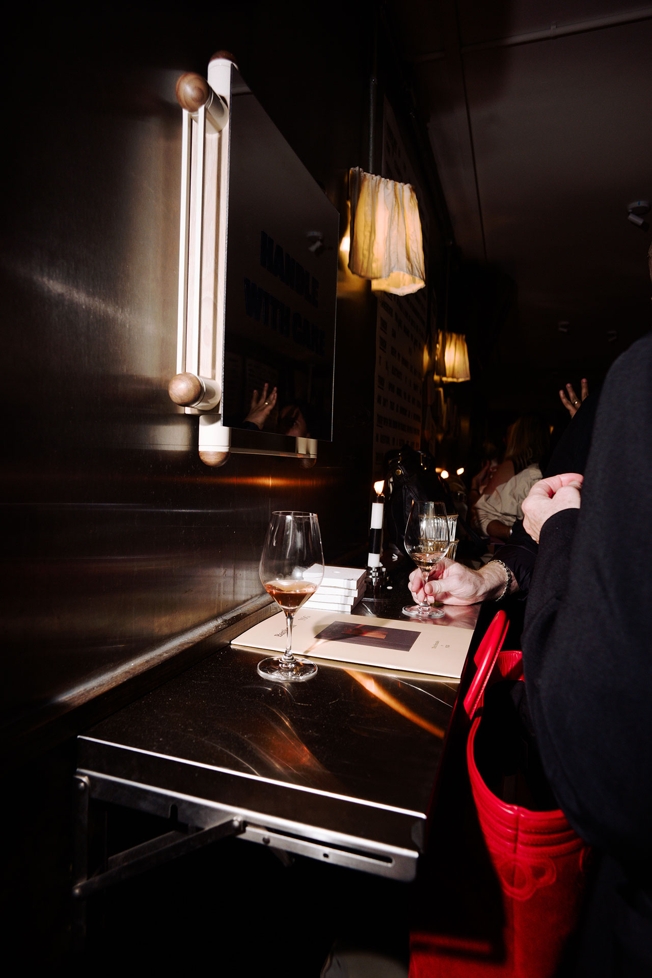 Two people stand at a metal counter in Bankston, with wine glasses, a candle, and a book. A mirror and hanging light fixtures add to the atmosphere in the dimly lit setting.