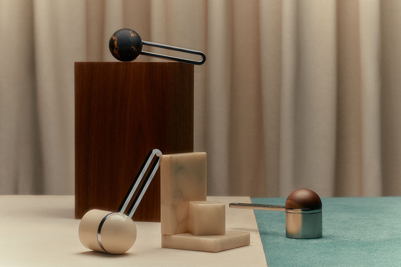 Three modern, sculptural coffee scoops with spherical handles are displayed on geometric blocks against a neutral draped background and a teal surface.