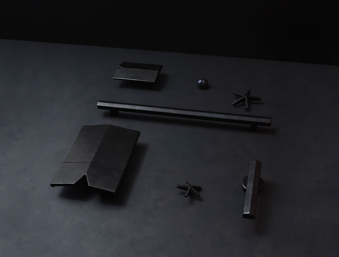 A set of matte black geometric objects, including bars, discs, and angular shapes, arranged on a dark, flat surface against a black background.