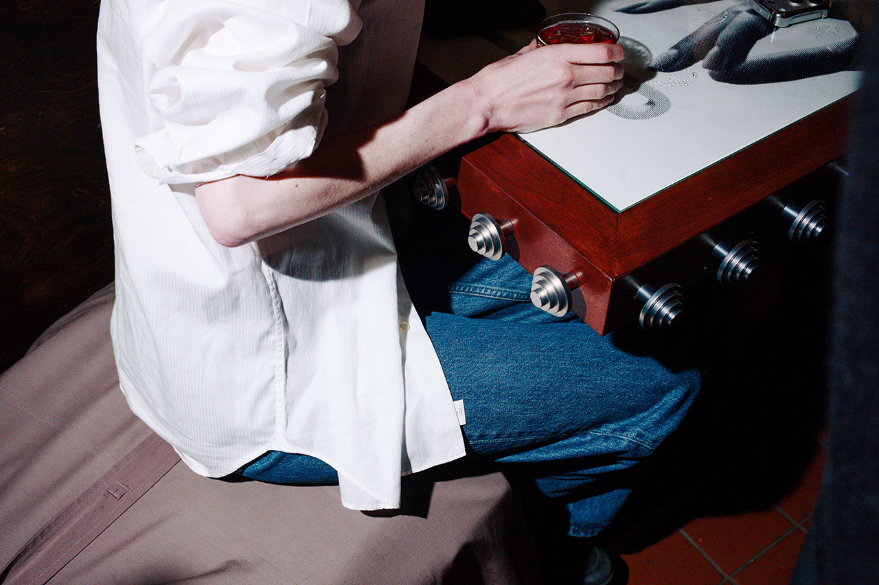 Bankston enthusiast in a white shirt and blue jeans sits at a foosball table, holding a drink in their right hand.
