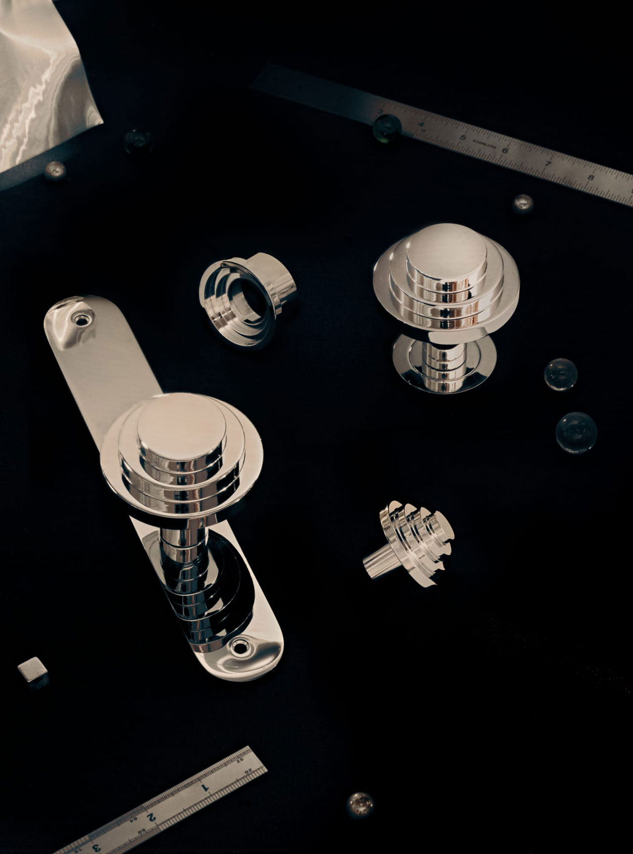 Chrome door handles and hardware parts are arranged on a dark surface alongside metal rulers and small spherical objects.