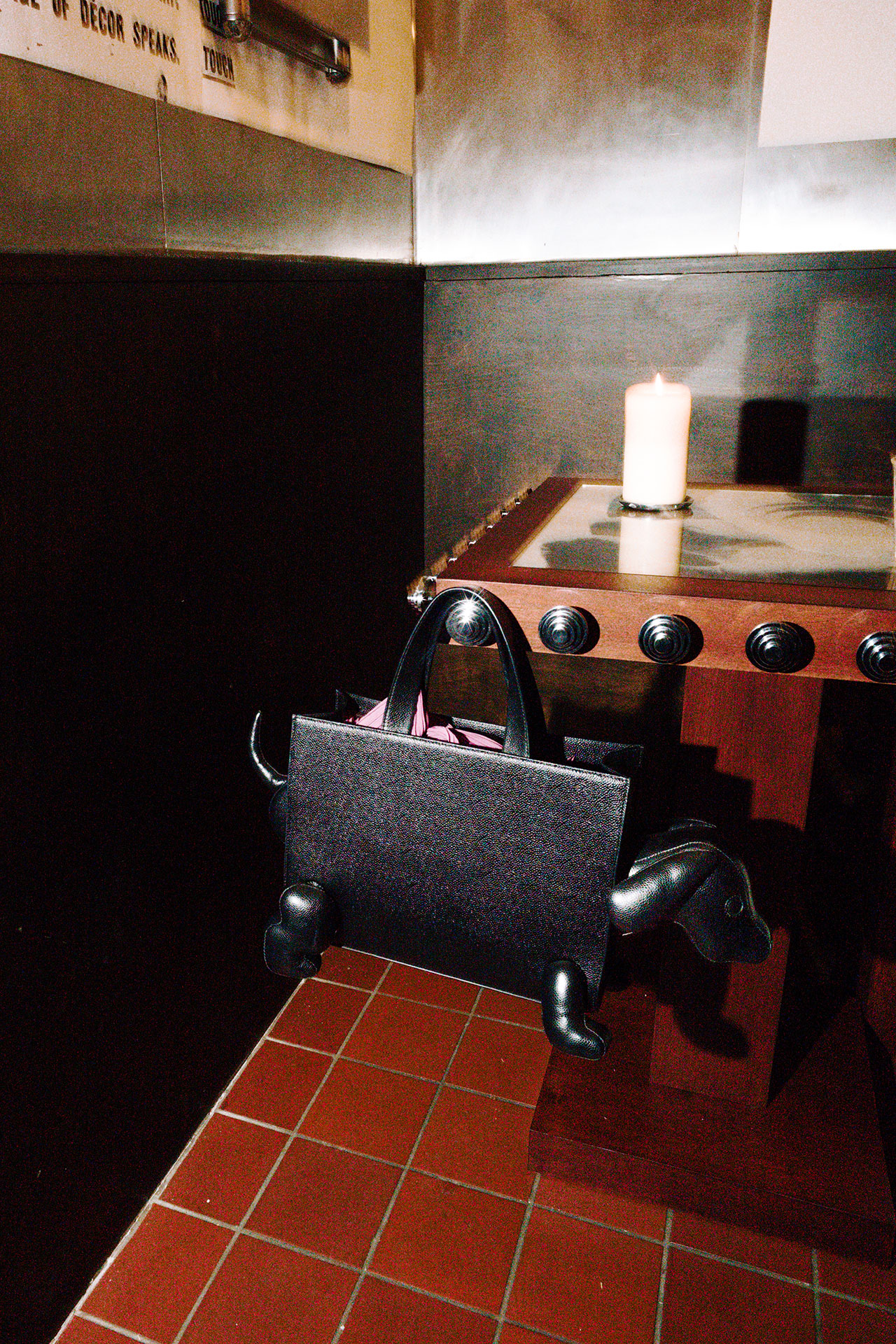 A black Bankston handbag with dog-shaped legs hangs from the edge of a wooden table, which has a lit candle on top; red tiled floor and black-and-white walls in the background.
