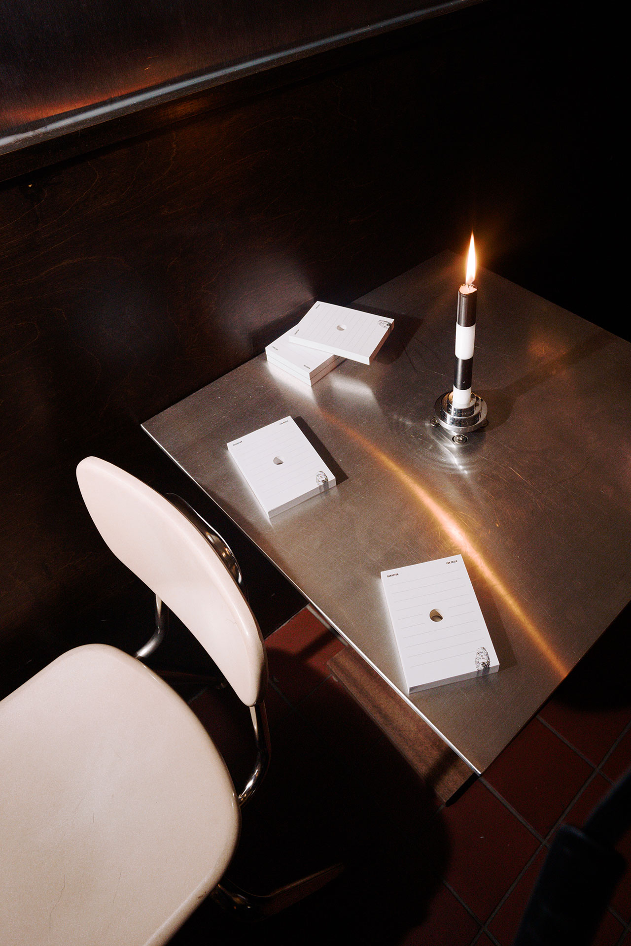 A Bankston metal table with a striped candle, three stacks of blank notepads, and a white chair sits in a dimly lit room.