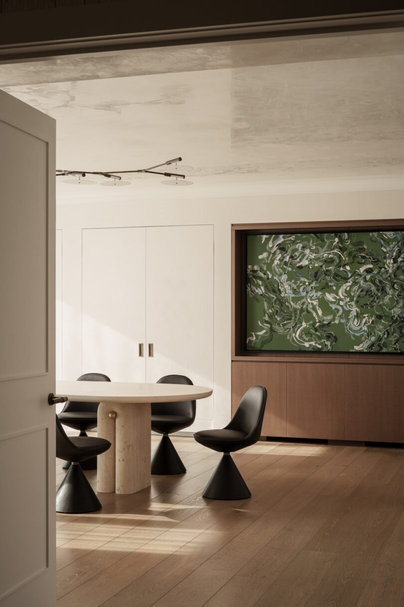 A modern dining area with a round table, four black chairs, light wood floors, white paneled walls, and a large abstract green artwork above a wooden cabinet.