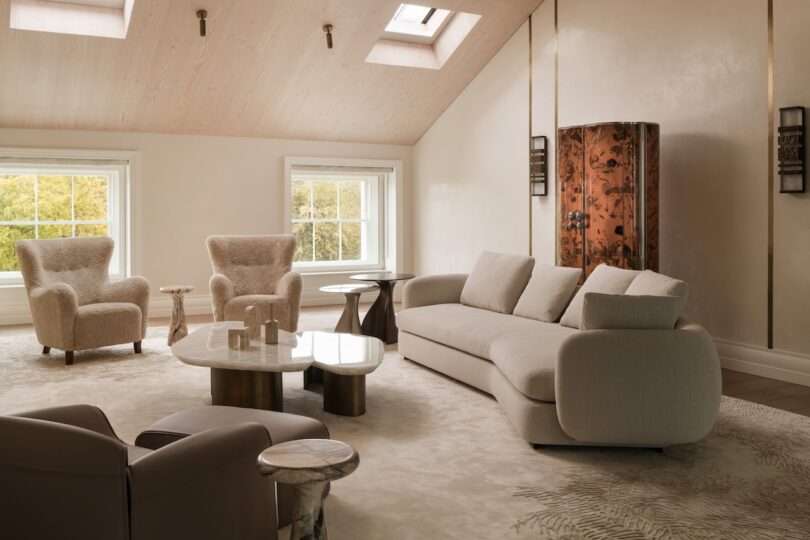 Bright, modern living room with a beige sofa, two textured armchairs, a marble coffee table, and skylights on a sloped wooden ceiling. A decorative cabinet stands by the wall.