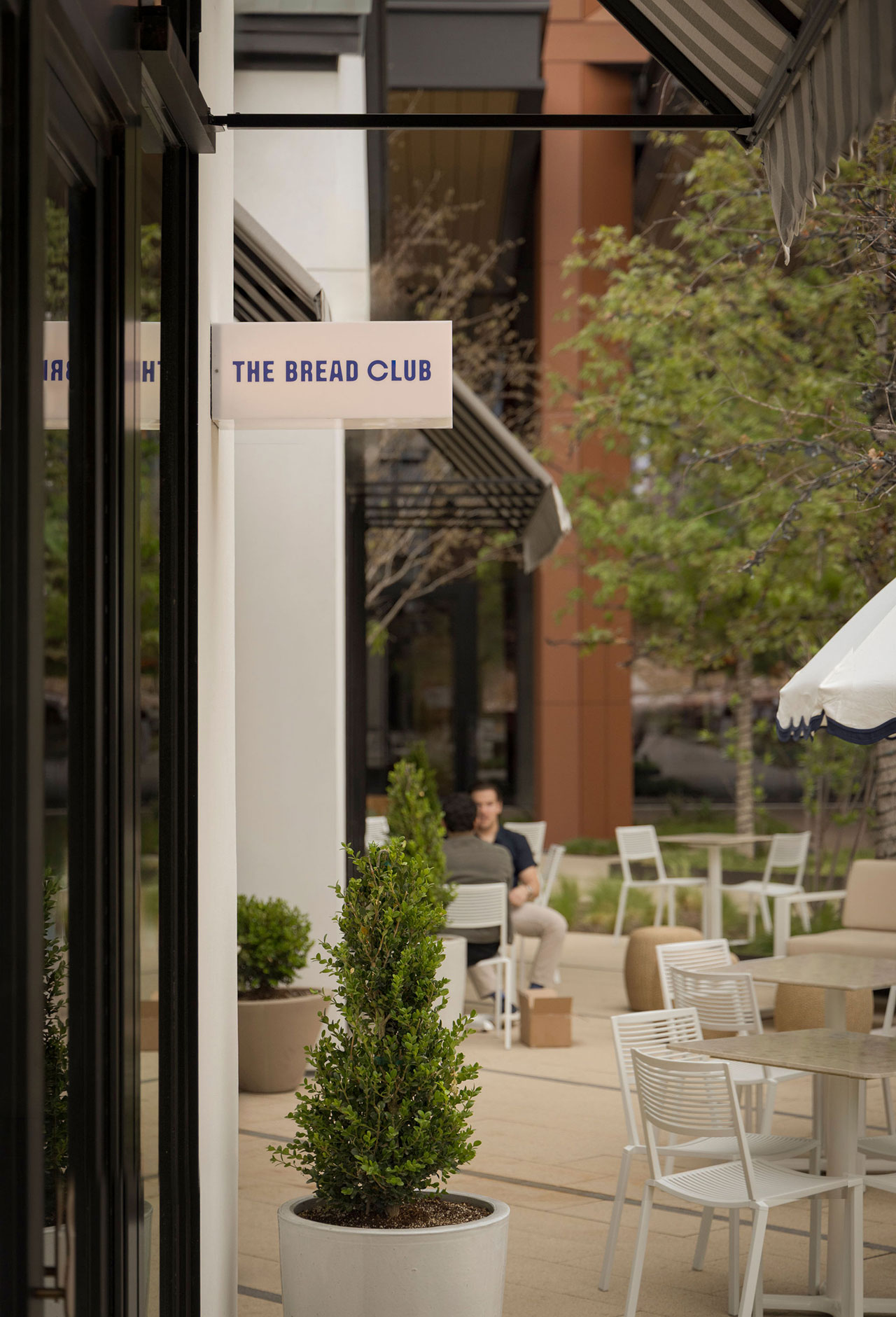 Outdoor seating area with white tables and chairs near potted plants, next to a building with a sign reading 