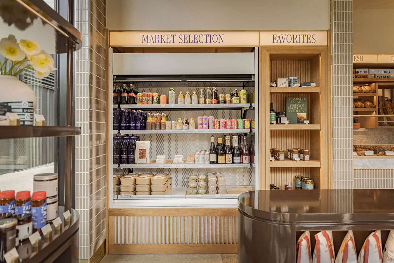 Refrigerated shelves in a store display bottled drinks, packaged foods, and jars. Adjacent open shelves hold more food items under signs reading 
