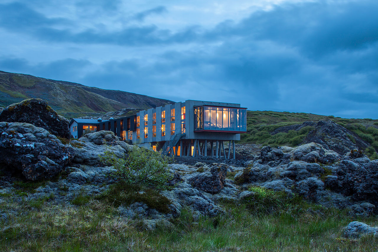 A modern building with large illuminated windows stands on stilts amid rocky terrain and sparse vegetation under a cloudy, twilight sky.