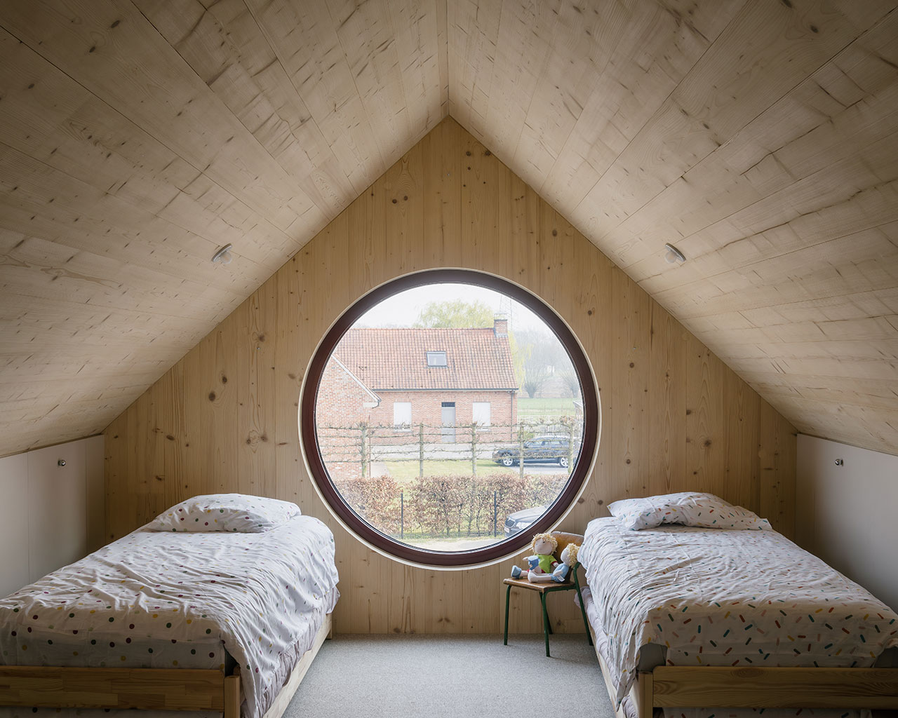 Two single beds with patterned bedding and soft felt accents are placed in a wooden attic room with a large round window overlooking houses outside.