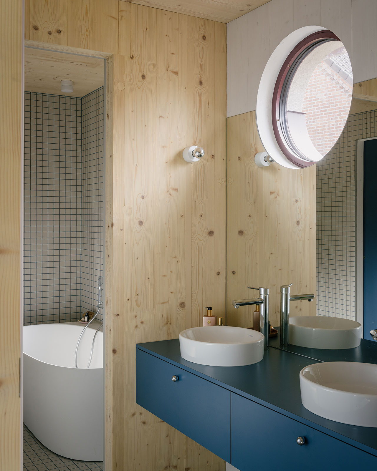 Modern bathroom with light wood walls and FELT accents, a round window, blue vanity with two vessel sinks, wall-mounted faucets, and a white freestanding bathtub in a tiled alcove.